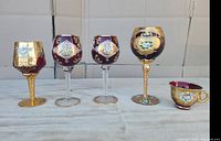 Set of five vintage Murano Venetian glasses in red and purple with 24K gold and enamel floral painting displayed on a light cloth background.
