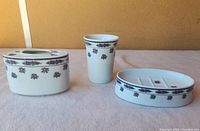 Porcelain bathroom set with soap dish, cup, and toothbrush holder displayed on a cloth surface against a plain background.