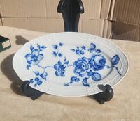 Oval white porcelain plate with blue rose floral decoration on black display stand on beige surface.