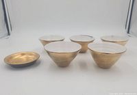 Front view of 5 gold and white Japanese pottery tea cups and 1 gold saucer arranged on white background, showing cups' shape and finish.