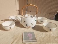 Porcelain teapot with bamboo handle and five matching teacups arranged with instruction booklet on fabric surface, cardboard background.