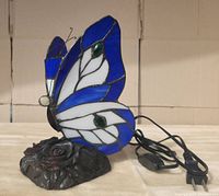 Side view of butterfly stained glass lamp showing the blue, white, and green glass wings and metal rose base with power cord.