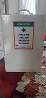 Closed white steel first aid cabinet with 'First Aid Station' label and carrying handle on top.