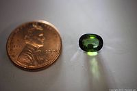 Photo showing the green tourmaline gemstone next to a penny for size comparison, highlighting the oval cut and vibrant green color.