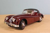 Close-up photo of die cast model Jaguar XK140 Drophead Coupe in burgundy with black convertible top on display surface.