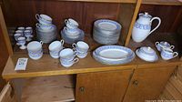 Photo showing the entire tea set displayed on a wooden cabinet shelf including plates, cups, saucers, egg cups, coffee pot, sugar bowl, cream pitcher, and platters, all featuring blue and gold pattern on white bone china.