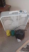 White Classic brand box fan with carry handle and power cord, an electric heater with control knobs, and two handheld flashlights (yellow and green) placed on carpeted floor.