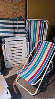 Four lounge chairs including two multi-color striped fabric metal framed chairs and two white resin slatted chairs stacked.