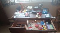 Wide view on wooden desk showing various office and stationery items both on top and in open drawers.
