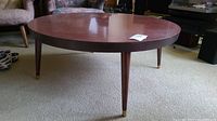 Side view showing the round coffee table with three tapered legs and brass feet, placed on carpeted floor.