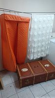 Photo showing orange clothes protector, sheer white curtains with circular accents, and vintage suitcase on the floor.