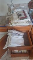 Folded assorted table linens and placemats stacked on a wooden table and inside an open drawer.