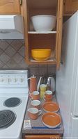 Full view of tea set on kitchen counter showing tall orange tea pot, six cups, two large bowls, and additional white and yellow bowls in cabinet above.