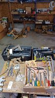 Wide shot of assorted hand tools laid out on a wooden bench, showing saw handles, pliers, scissors, screwdrivers, clamp, and hardware boxes in the background