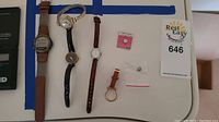 Five wristwatches laid out flat with some small accessories on a white surface.