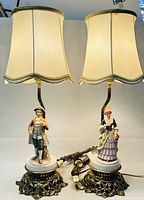 Pair of vintage figural table lamps with cream scalloped fabric shades trimmed in gold, featuring porcelain figurines of a man and woman on marble bases with ornate metal stands viewed from front.