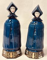 Two ceramic Mulher do Capote liquor bottles shaped as ladies, standing side by side showing front view, blue cloaks, detailed faces, hexagonal bases with gold and white text.
