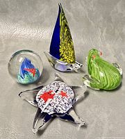 Photo showing all four paperweights: sailboat, star-shaped, ball with fish, and green frog