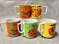 Photo showing all five bright colorful Nasco mugs with stylized fruit graphics and white handles arranged in two rows