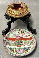 Photo showing ornate copper ashtray on black plastic dragon stand with hand-painted ceramic ashtray in front.