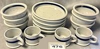 Photo showing 5 stacked 10 inch plates, 6 stacked bowls, and 4 mugs in front, all featuring cream colored ceramic with a blue band near the rims.