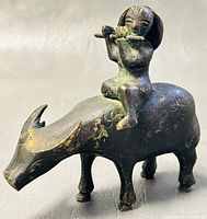 Side view of the cast bronze figurine showing the person playing flute seated on water buffalo with dark finish and patina.