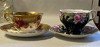 Two complete teacup and saucer sets, one with black floral design and one with gold rose floral design