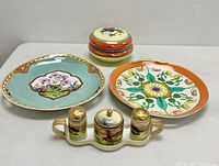 Wide shot showing all items: two decorative plates with floral patterns and a porcelain condiment set with holder including salt, pepper, and mustard pot.