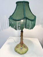 Porcelain table lamp with green fabric shade and fringed trim on a base shaped like a tree trunk with a small gnome figure seated on grassy ground.