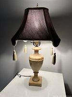 Full view of the lit lamp showing fabric shade with tassels and alabaster marble urn-shaped base on square pedestal.