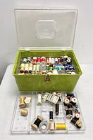 Open green plastic storage case filled with multiple wooden and plastic spools of thread of various colors.