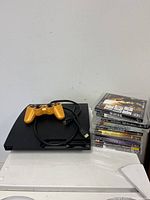 Black Sony PlayStation 3 console with orange controller and HDMI cable, accompanied by stacked game cases.