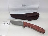 Knife with polished carbon steel blade and reddish maple handle resting on white surface with typed letter and brown leather sheath behind it.