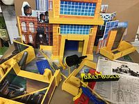 Playset parts spread around a yellow building facade with a blue window section. Various assembly components and accessories including black cannon, flexible hoses, and red structural pieces visible.