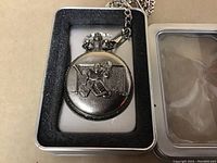 Closed view of silver-tone pocket watch featuring an embossed hockey goalie in front of goal net on the cover, inside foam case insert and silver chain visible.