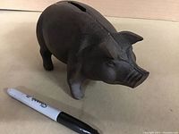 View of dark cast iron piggy bank with coin slot on top, placed next to a Sharpie marker for size reference.