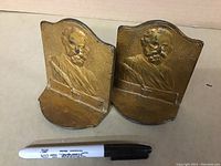 Front view of two antique cast iron bookends depicting Henry Wadsworth Longfellow busts with bronze finish and textured backgrounds, with a Sharpie pen for scale.