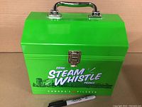 Front view of a bright green metal lunch box with a sturdy chrome handle on top and a latch in front. Bold white text reads 'Steam Whistle' with smaller text 'Drink' above and 'Pilsner' below. Advertisement includes 'Canada's Pilsner' text with small red icon.