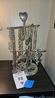 Full view of several silver tone necklaces and bracelets hanging on an ornate silver tone jewelry holder with heart-shaped finial.
