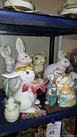Shelf display of Easter-themed decor including ceramic rabbits, a large white rabbit with yellow flower hat, and smaller figurines