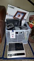 Plastic basket full of electronics including Coby digital picture frame in box, Kodak camera, and Sony cassette player below basket.