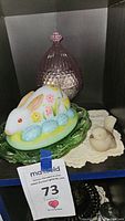 Pink depression glass lidded jar with hobnail pattern and bird figurine lid, next to a hand-painted rabbit candy dish with blue eggs on a green plate and ceramic bird figurine on a lace doily.