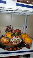 Photo showing multiple ceramic Halloween items on shelf including 3 lidded pumpkin jars, Halloween plates, and other decorations