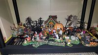Wide view of multiple Halloween-themed figurines and decorations including haunted houses, witches, ghosts, and spooky trees arranged on a black surface.