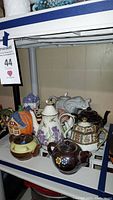 Shelf display of multiple assorted ceramic teapots including decorative shapes and floral designs, showing front view with elephant and pumpkin house figurines
