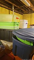 Stacked storage totes in a basement area showing various sizes and colors including blue, green, gray, clear, and white totes. Some lids present, others missing.