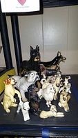 Front view of 14 vintage ceramic dog figurines in various breeds and poses on a dark surface under lighting.