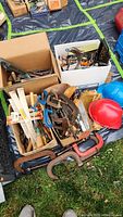 Overview of multiple cardboard boxes with various hand tools, clamps, files, red and blue hard hats, and tool holders on ground with tarp underneath