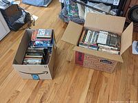 View of two open cardboard boxes on wooden floor filled with DVDs, showing the assortment and quantity for the lot.