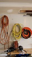 Orange, yellow, and black extension cords hanging on garage pegboard wall with two battery chargers below